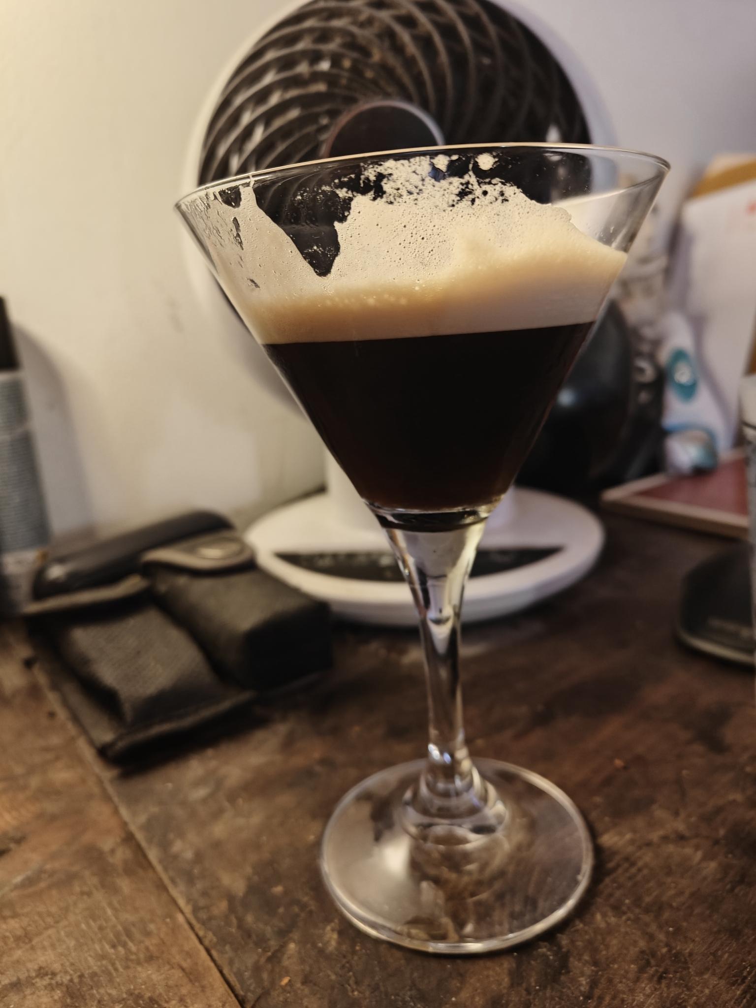 Un verre d'expresso martini posé sur un meuble en bois devant un petit ventilateur boule.