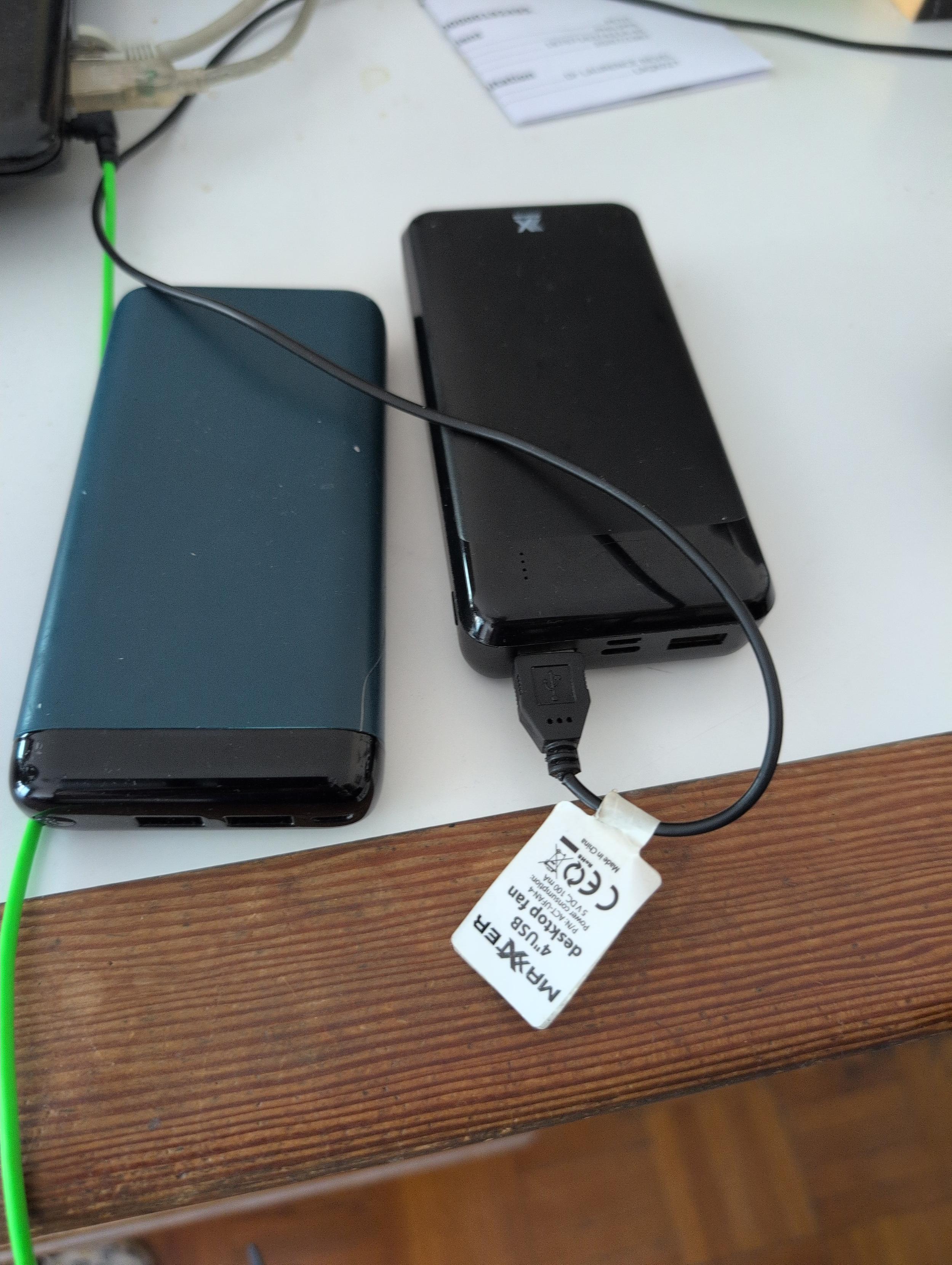 Sur un plateau de bureau blanc à bordure bois clair, deux batteries rechargeables (powerbank). Le bleu à gauche a une capacité de 10.000 mAh et le noir à droite 20.000 mAh.