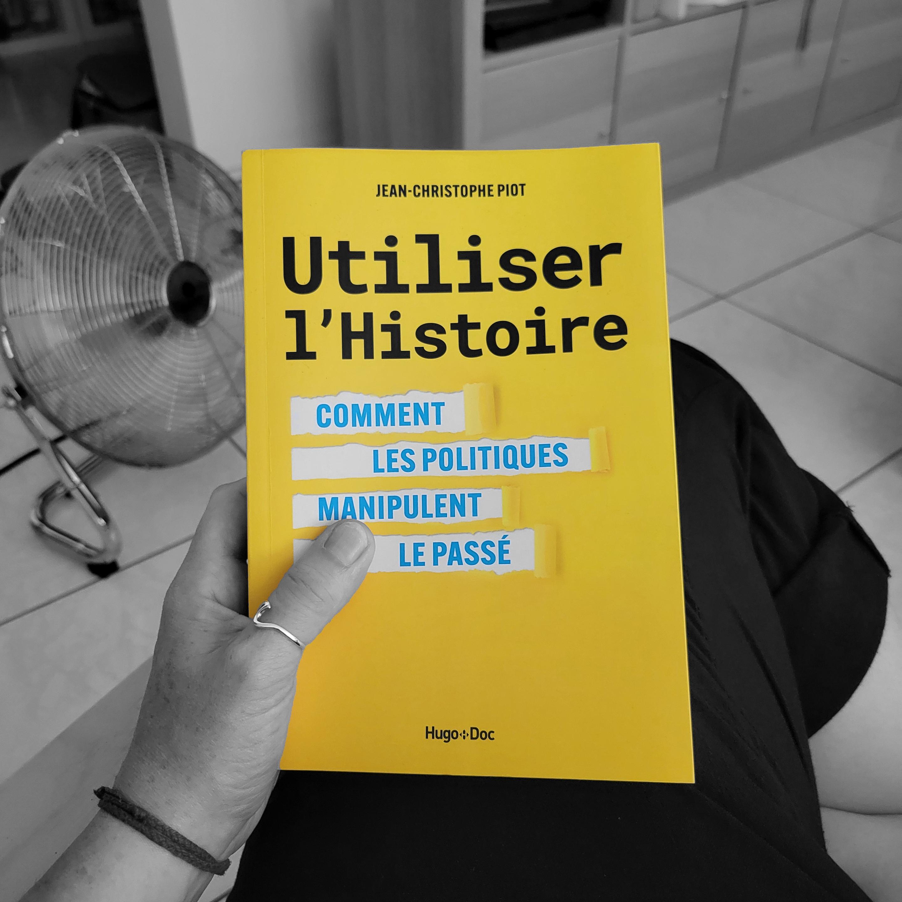 Photo en couleurs sélectives de ma main tenant le livre Utiliser l’Histoire. La couverture est jaune vif. On distingue un ventilateur sur la gauche en arrière-plan.